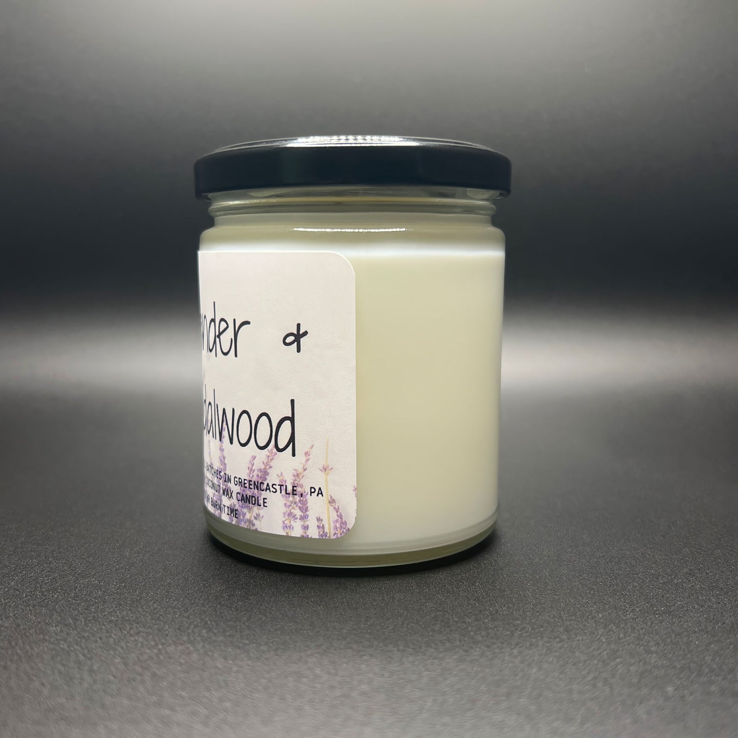 Angled view of a closed Lavender and Sandalwood candle by Elysium Candle Co. The image displays the candle’s glass jar, black lid, and white label adorned with lavender illustrations and product details, highlighting the 40-50 hour burn time and GreenCastle, PA origin.