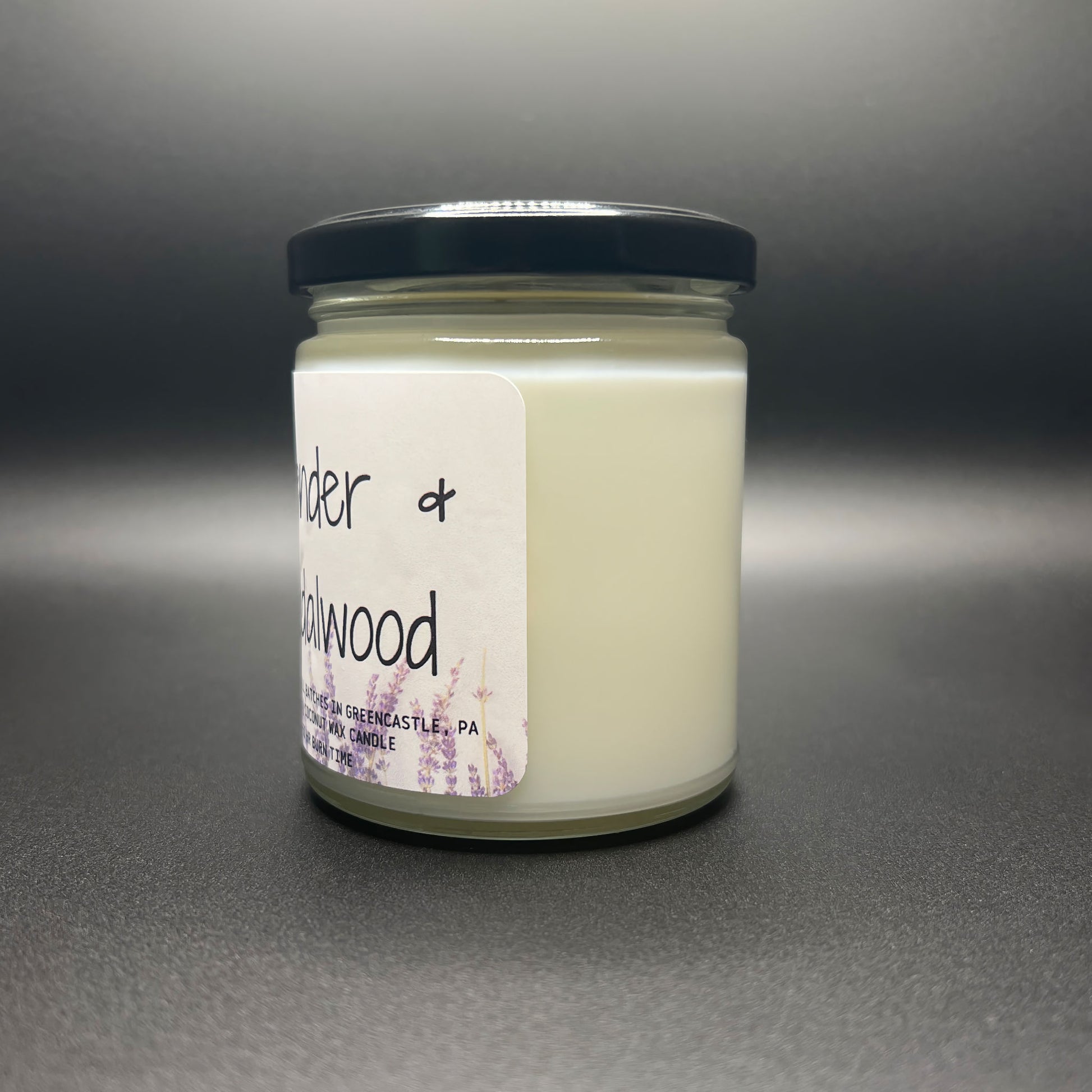 Angled view of a closed Lavender and Sandalwood candle by Elysium Candle Co. The image displays the candle’s glass jar, black lid, and white label adorned with lavender illustrations and product details, highlighting the 40-50 hour burn time and GreenCastle, PA origin.