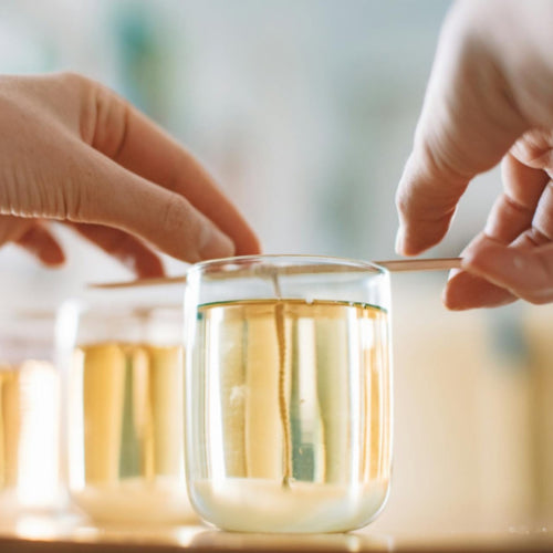 Hands carefully aligning the wick in a transparent glass jar filled with golden melted coconut soy wax, illustrating the meticulous process of making handmade, non-toxic candles.