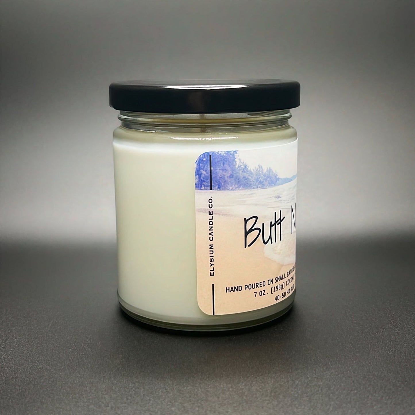 A side view of Elysium Candle Co.’s Butt Naked candle, displaying a smooth coconut soy blend wax and a label with a peaceful beach imagery.