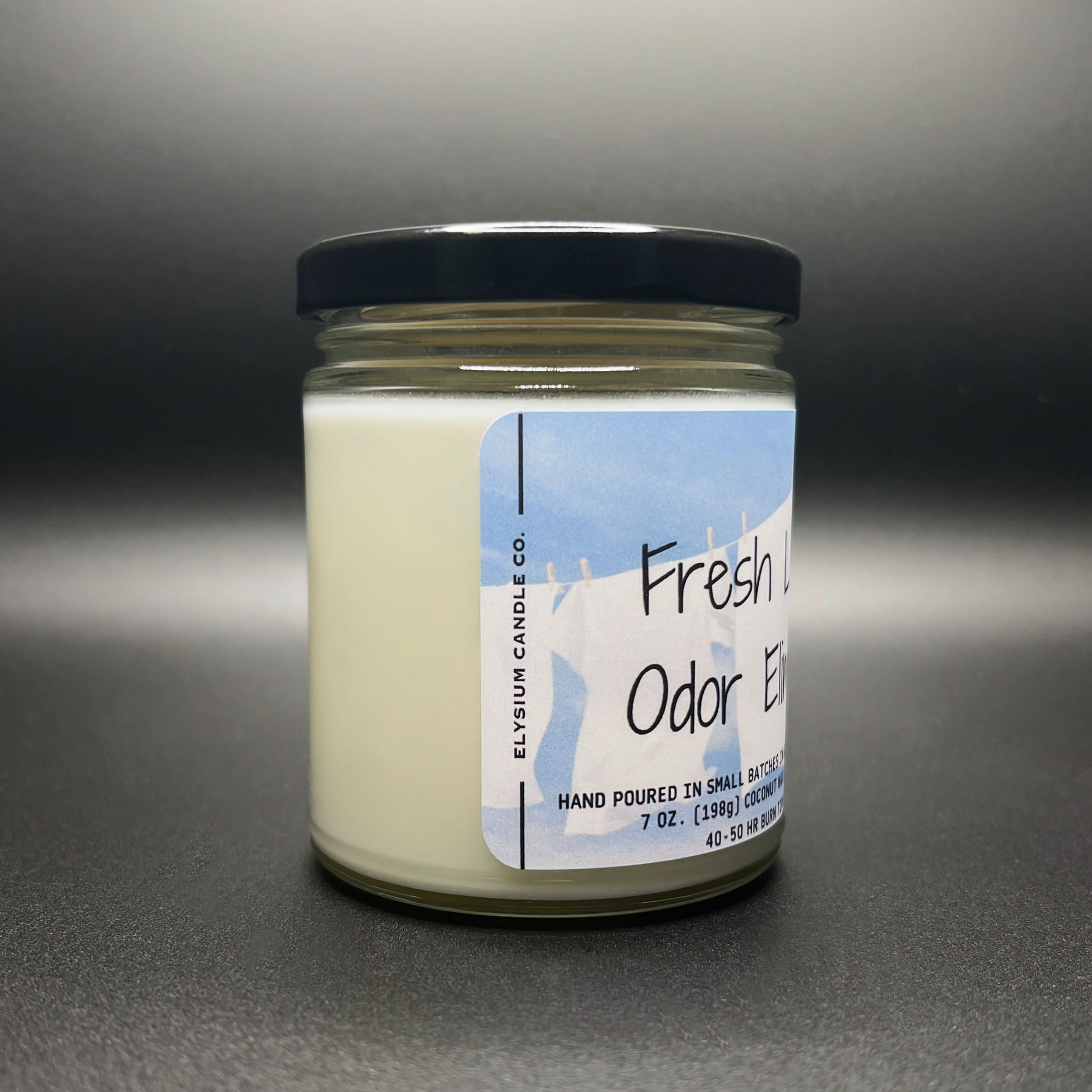 Side view of a ‘Fresh Linen Odor Eliminator’ candle, featuring a clear jar with a black lid, and a label with blue sky and laundry imagery indicating the scent.