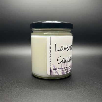 A Lavender and Sandalwood scented coconut-soy blend candle by Elysium Candle Co. presented against a dark background. Side view showing the creamy white wax in a clear glass jar with a black lid and a label that features the company name and scent details. The label has a minimalistic design with lavender sprig accents.