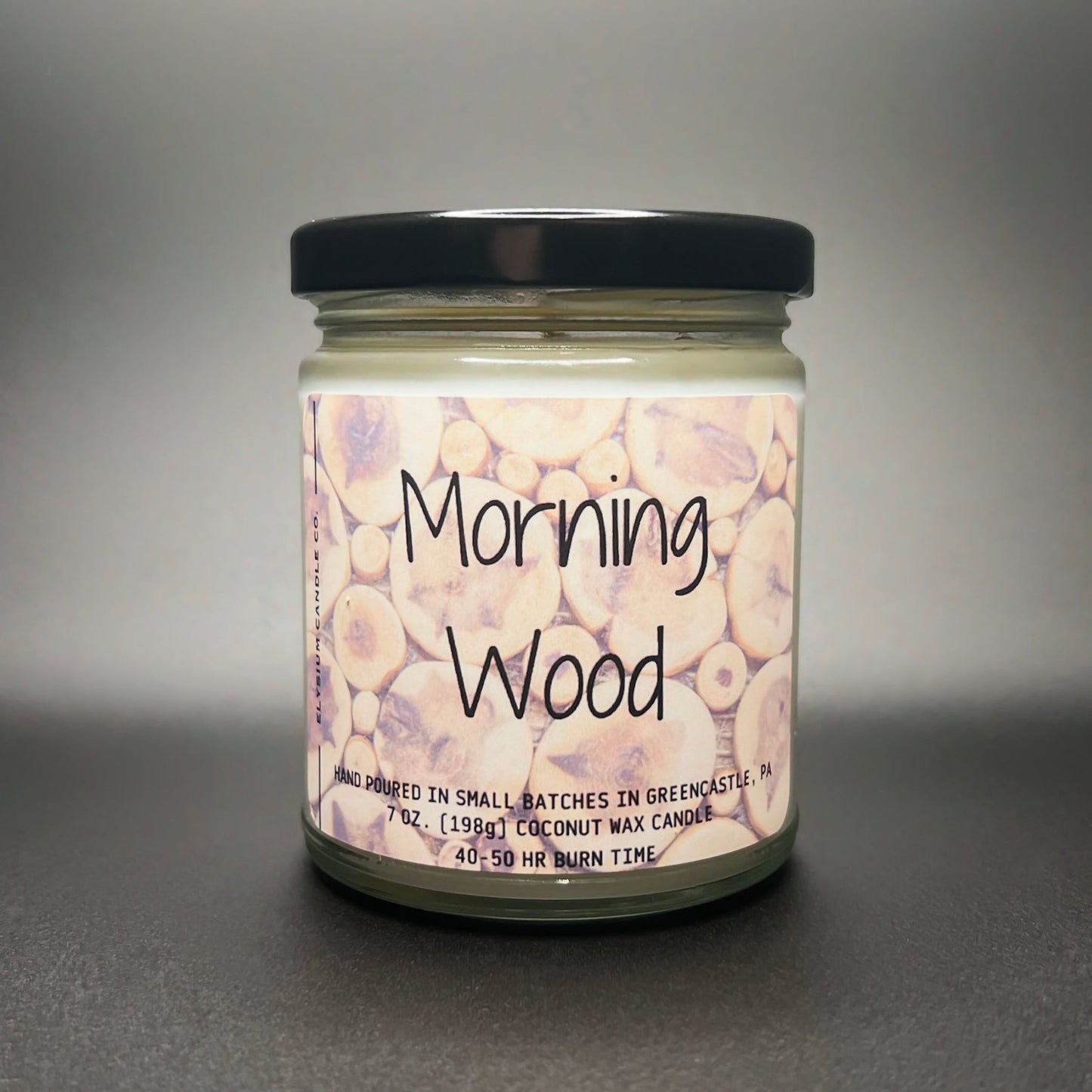 A 7 oz coconut soy wax candle named ‘Morning Wood’ by Elysium Candle Co., featuring a wood log pattern label, hand-poured in Greencastle, PA with a 40-50 hour burn time.