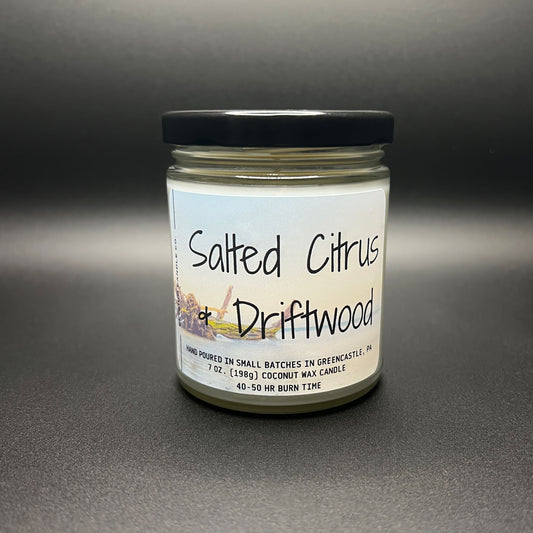 Front view of a Salted Citrus and Driftwood coconut wax candle by Elysium Candle Co., with a crisp, clear label featuring a beach scene. The jar is topped with a sleek black lid, and the creamy white wax is visible through the glass.