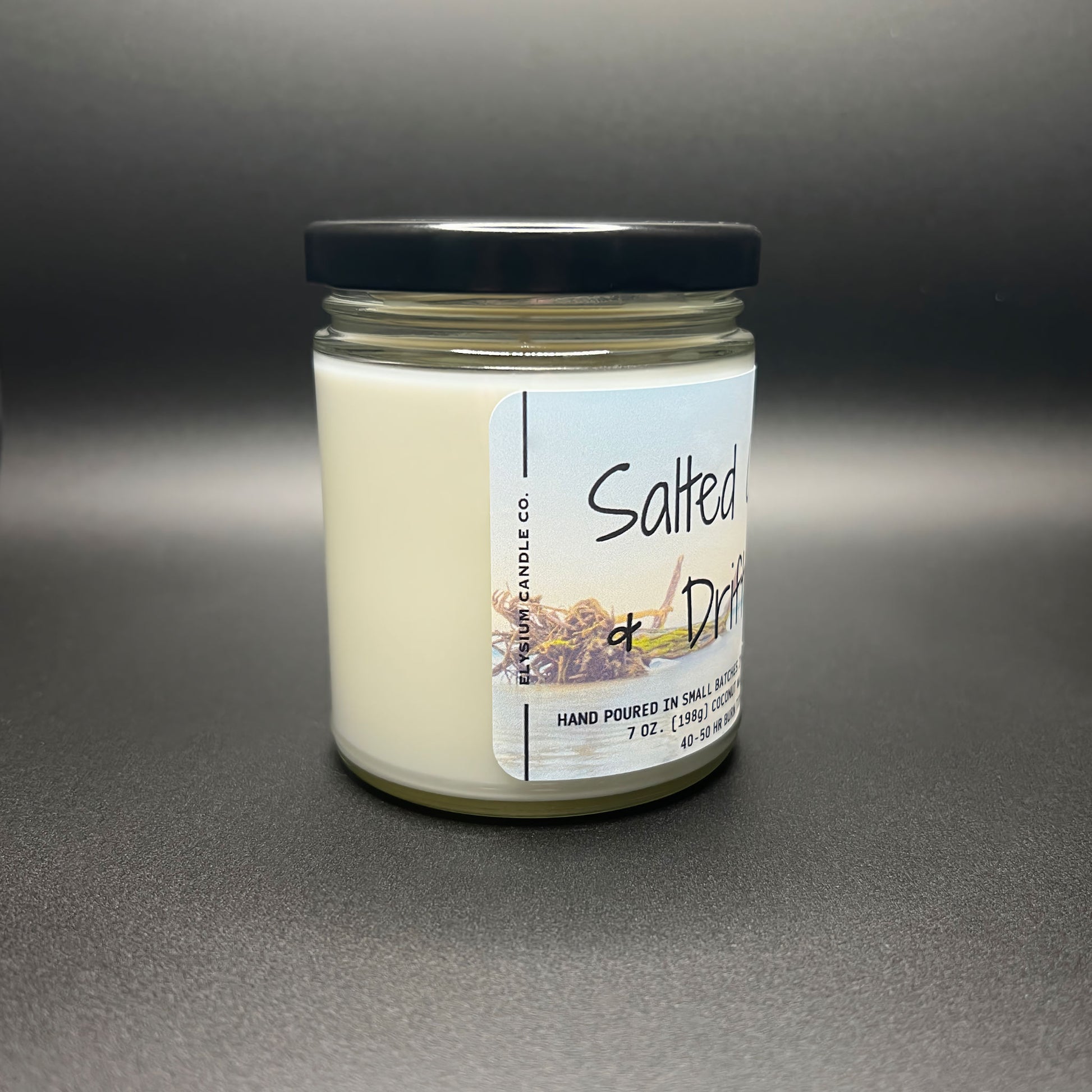 Side view of Elysium Candle Co.’s Salted Citrus and Driftwood candle, highlighting the creamy texture of the coconut wax inside a transparent jar with a black lid. The label displays the hand-poured and small batch details.